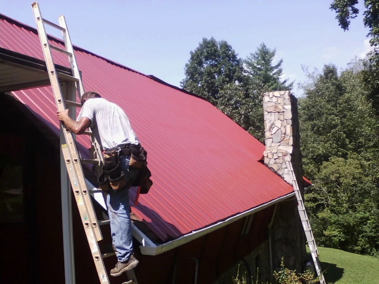 Roofer on a ladder beside a red metal roof and stone chimney — Soffit & Fascia Repair by Roofing Brothers in Economy